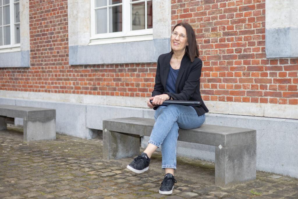 Wendy zit op een bankje buiten met de laptop op de schoot en kijkt recht in de camera